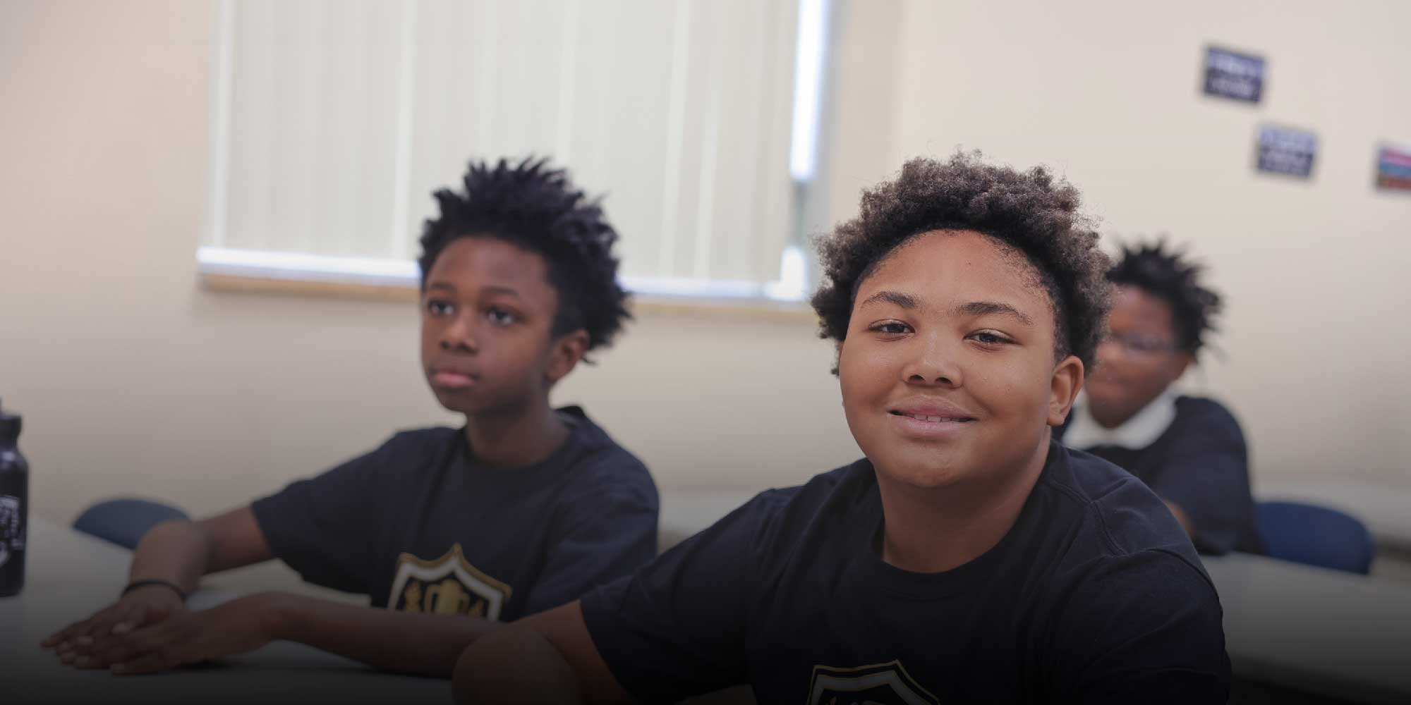 Students in school T-shirts seated in a classroom, one smiling at the camera.