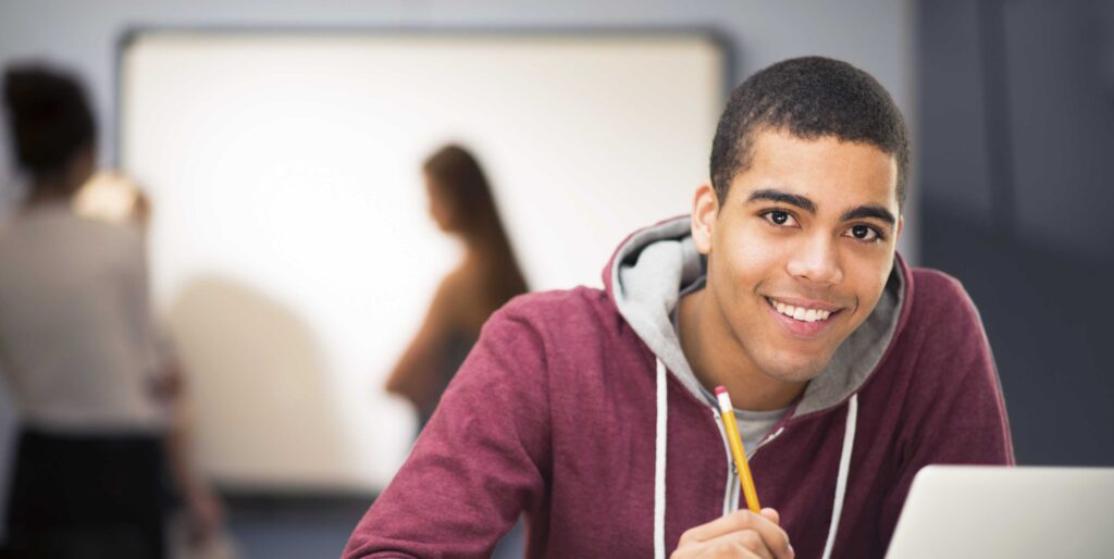 Smiling high school student holding a pencil at a laptop in a classroom